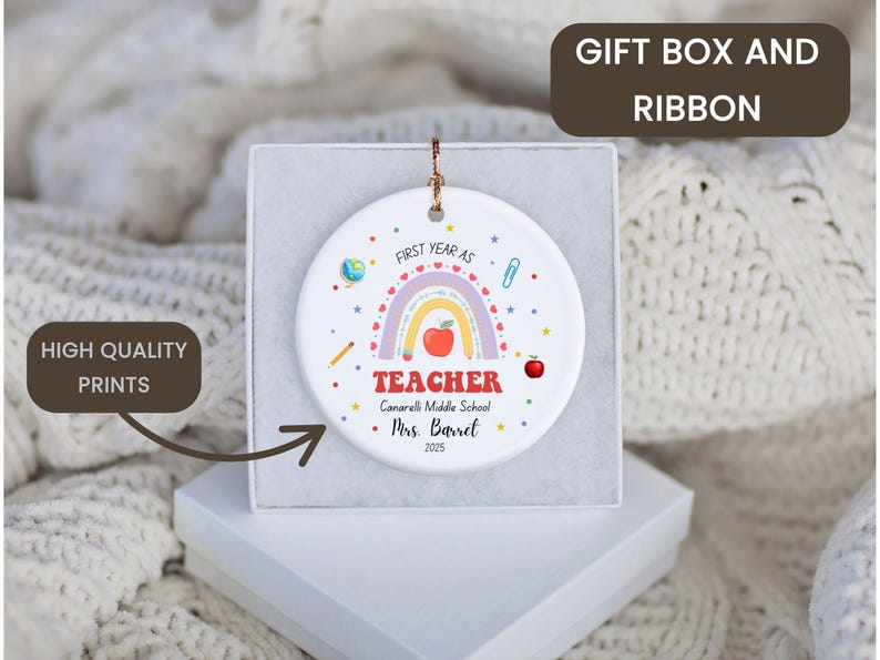 Peut inclure: Ornement en c&eacute;ramique blanc avec un arc-en-ciel, une pomme et le texte "FIRST YEAR AS TEACHER". L'ornement est pr&eacute;sent&eacute; dans un coffret cadeau blanc. Le texte comprend &eacute;galement "Canarelli Middle School" et "Mrs. Barret 2025."