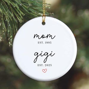 Puede incluir: Adorno navideño redondo de cerámica blanca con las palabras en negro "mom EST. 1995" y "gigi EST. 2025". Un pequeño corazón rojo en la parte inferior. El adorno cuelga de un hilo dorado.