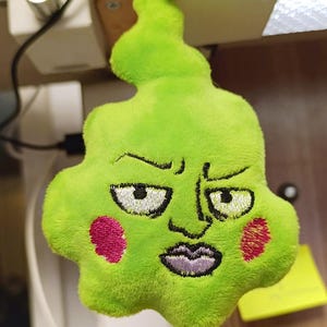 Op de afbeelding: Groene pluche sleutelhanger met een cartoon gezicht. Het gezicht heeft een fronsende uitdrukking met roze wangen en een paarse mond.