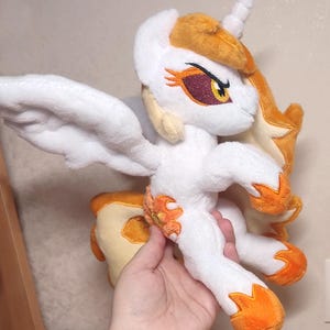 Könnte beinhalten: Ein Plüschtier eines weißen Einhorns mit orangefarbenen und gelben Akzenten. Das Einhorn hat ein weißes Horn, Flügel und eine fließende Mähne. Die Augen sind lila und gelb. Das Plüschtier ist weich und detailliert, mit gestickten Details.