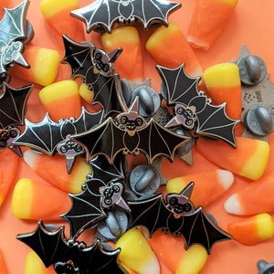 May include: A close-up of a group of black enamel pins shaped like bats with purple accents. The pins are scattered on a surface with orange and yellow candy corn.