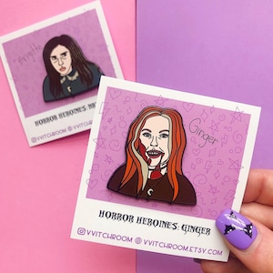 May include: Two enamel pins featuring horror movie characters. The first pin is of a woman with long dark hair and the text "Brigitte". The second pin is of a woman with long red hair and the text "Ginger". Both pins have a pink background with black and white stars, hearts, and lightning bolts.