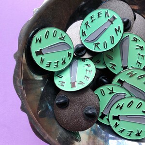 May include: A collection of green enamel pins with a black outline. Each pin features a machete and the text "Green Room" in a circular design.