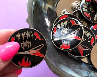 Op de afbeelding: Een close-up van een zwarte en rode emaille pin met een gitaar en vlammen. De pin heeft de tekst "The Devil's Candy" in gouden letters.