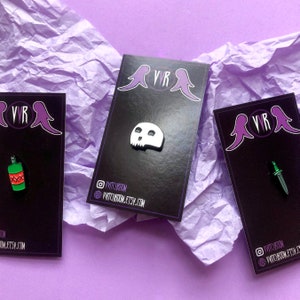May include: Three black cards with white enamel pins. The pins are a skull, a bottle of alcohol, and a dagger. The cards have the text "VVR" and the social media handle "@VVITCHROOM".