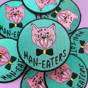 May include: A set of nine teal and pink embroidered iron-on patches featuring a cartoon cat with a bow tie and the text "MAN-EATERS".