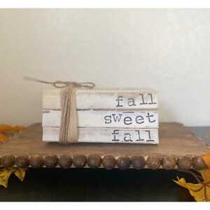 Puede incluir: Una pila de tres libros pintados de blanco con las palabras "fall sweet fall" impresas en ellos. Los libros están atados con cuerda y están sobre una bandeja marrón con un borde de cuentas. Hay hojas de otoño a cada lado de la bandeja.