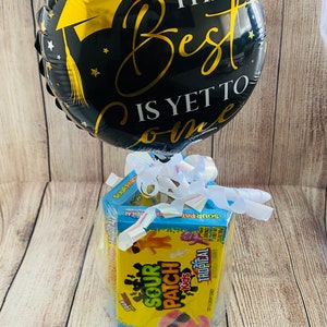 May include: A black and gold graduation balloon with the text "The Best is Yet to Come" and a graduation cap. The balloon is tied to a box of Sour Patch Kids Tropical candy.