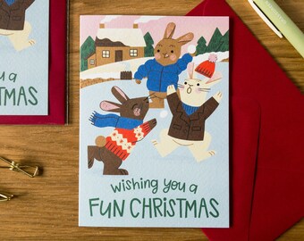 May include: Christmas greeting cards featuring cartoon rabbits playing in the snow. The cards have a light blue background with the text "Wishing you a FUN CHRISTMAS" and "Wishing you a CHRISTMAS". The cards are paired with red envelopes.