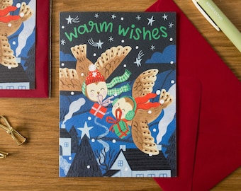 May include: Holiday greeting card with a whimsical illustration of owls flying in a snowy night scene. The card features the text "warm wishes" in green. A red envelope and a green pen are also visible.