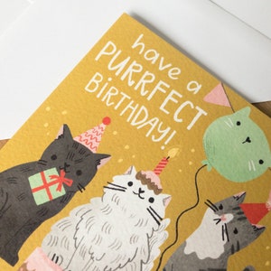 May include: A yellow birthday card with the text "have a purrfect birthday!" and three cats wearing party hats. One cat is holding a gift, another is wearing a cake hat with a candle, and the third is holding a green balloon.
