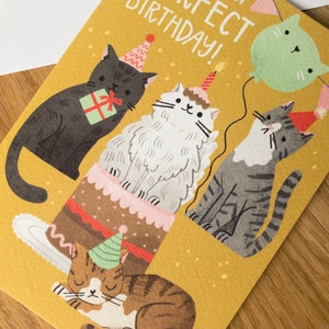 May include: A yellow birthday card with a cat-themed design. Four cats are depicted, one wearing a party hat and sitting on a cake, another holding a gift, and two others wearing party hats. The card reads "Happy Purrfect Birthday!"