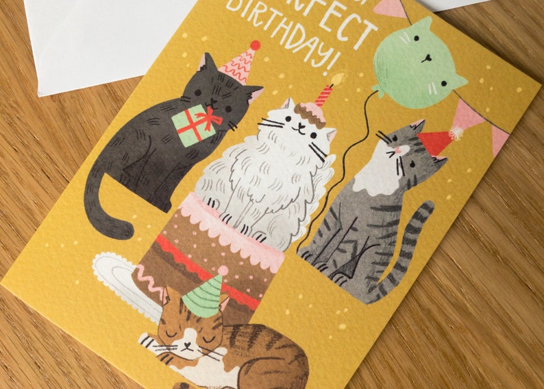 May include: A yellow birthday card with a cat-themed design. Four cats are depicted, one wearing a party hat and sitting on a cake, another holding a gift, and two others wearing party hats. The card reads "Happy Purrfect Birthday!"