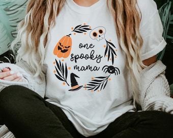 May include: White t-shirt with a black and white graphic design of a wreath with a pumpkin, skull, spider, witch hat, and the text "one spooky mama".