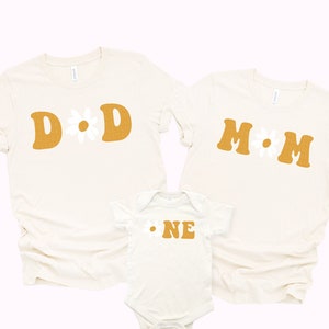 May include: Three cream-colored t-shirts with a retro daisy design. The shirts say "DAD", "MOM", and "ONE" in a mustard yellow color.