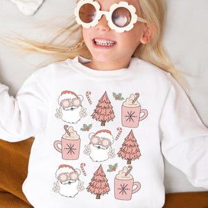 May include: White sweatshirt featuring a repeating pattern of Santa Claus faces, pink Christmas trees, and mugs of hot cocoa. The Santa Claus illustrations are wearing sunglasses and giving the peace sign. The sweatshirt is a festive holiday design.