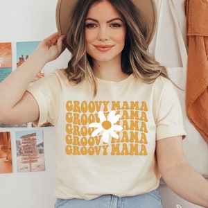 May include: A light yellow t-shirt with the words "GROOVY MAMA" in orange, stacked vertically with a white daisy in the center. The shirt is being worn by a person. The person is wearing a brown hat and blue jeans.