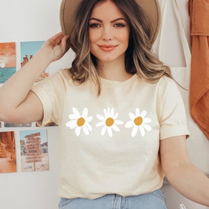 K&ouml;nnte beinhalten: Eine Frau tr&auml;gt ein hellgelbes T-Shirt mit drei wei&szlig;en G&auml;nsebl&uuml;mchen mit gelben Zentren. Die G&auml;nsebl&uuml;mchen sind in einer Reihe &uuml;ber die Mitte des Shirts angeordnet.