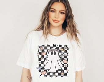 May include: White t-shirt with a black and white chequered design featuring a ghost with a flower crown, bats, and pumpkins.