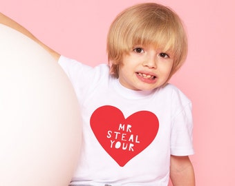 May include: A white t-shirt with a large red heart graphic that says "MR STEAL YOUR" in white text. The shirt is worn by a child, who is sitting next to a large white ball. The background is a solid pink.