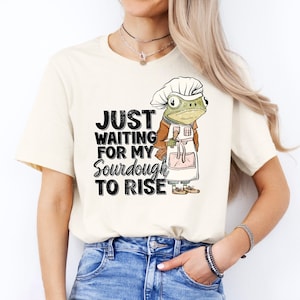 May include: A white t-shirt with a graphic of a frog wearing a chef's hat and apron. The text on the shirt reads "Just waiting for my sourdough to rise."