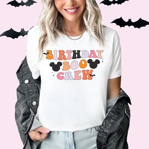 Puede incluir: Camiseta blanca con el texto "Birthday Boo Crew" en letras retro de colores. El texto est&aacute; decorado con murci&eacute;lagos negros y orejas de Mickey Mouse.