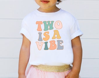 May include: A white t-shirt with the text "TWO IS A VIBE" in colorful, retro-style letters. The letters are in shades of pink, orange, yellow, green, and blue.