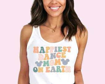 以下が含まれることがあります： カラフルなレトロフォントで「Happiest Dance Mom on Earth」と書かれた白いタンクトップ。デザインにはミッキーマウスのシルエットが含まれています。女性は青いデニムショーツを着用しています。