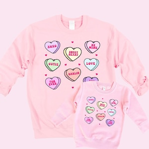May include: Two pink sweatshirts with a Valentine's Day theme. The larger sweatshirt has a pattern of candy hearts with phrases like "XOXO", "Sweet Heart", "Be Mine", "Gutie", "Love", "Darlin", "For Ever". The smaller sweatshirt has the same pattern but with fewer hearts.