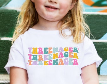 May include: A white t-shirt with the text "THREEENAGER" repeated three times in a rainbow of colors. The text is in a retro font style.