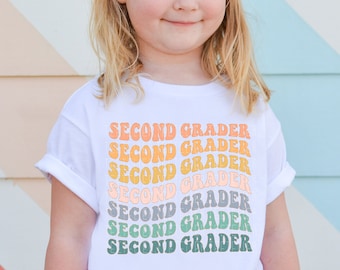 Könnte beinhalten: Ein weißes T-Shirt mit dem Text "SECOND GRADER", der in einem Regenbogenmuster wiederholt wird. Der Text ist in einem Retro-Schriftart-Stil.