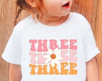May include: A white t-shirt with the word "THREE" repeated three times in pink, orange, and white with a white daisy in the center. The daisy has a brown center.