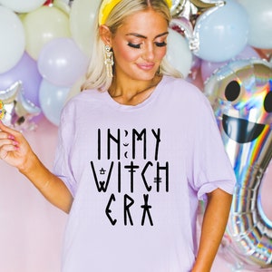 May include: A light purple t-shirt with the text "IN MY WITCH ERA" in black. The shirt is worn by a person with blonde hair, a yellow headband, and skeleton earrings. Balloons in pastel colors are in the background.
