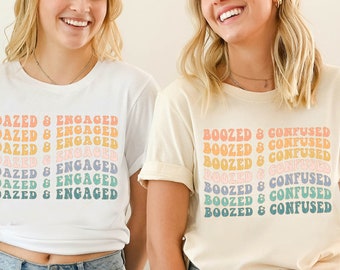 May include: Two women wearing t-shirts with different slogans. The woman on the left is wearing a white t-shirt with the text "Dazed & Engaged" repeated in rainbow colours. The woman on the right is wearing a cream t-shirt with the text "Boozed & Confused" repeated in rainbow colours. Both women are wearing blue jeans.