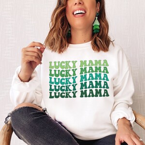May include: White sweatshirt with the text "Lucky Mama" repeated five times in a green and blue gradient. The text is in a retro font style.