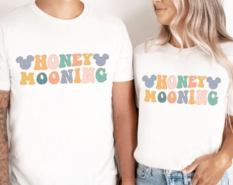 May include: Two white t-shirts with the text "Honeymooning" in colourful retro font with Mickey Mouse ears on either side of the text.