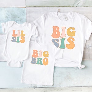 May include: A white baby bodysuit with the text LIL SIS, a white t-shirt with the text BIG BRO, and a white t-shirt with the text BIG SIS. Each garment features colorful, retro-style lettering in shades of orange, pink, blue, and green.