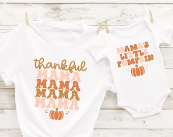 May include: A white t-shirt with the text "thankful mama" repeated four times in a stacked design with a pumpkin graphic. A white bodysuit with the text "mama's little pumpkin" and a pumpkin graphic.