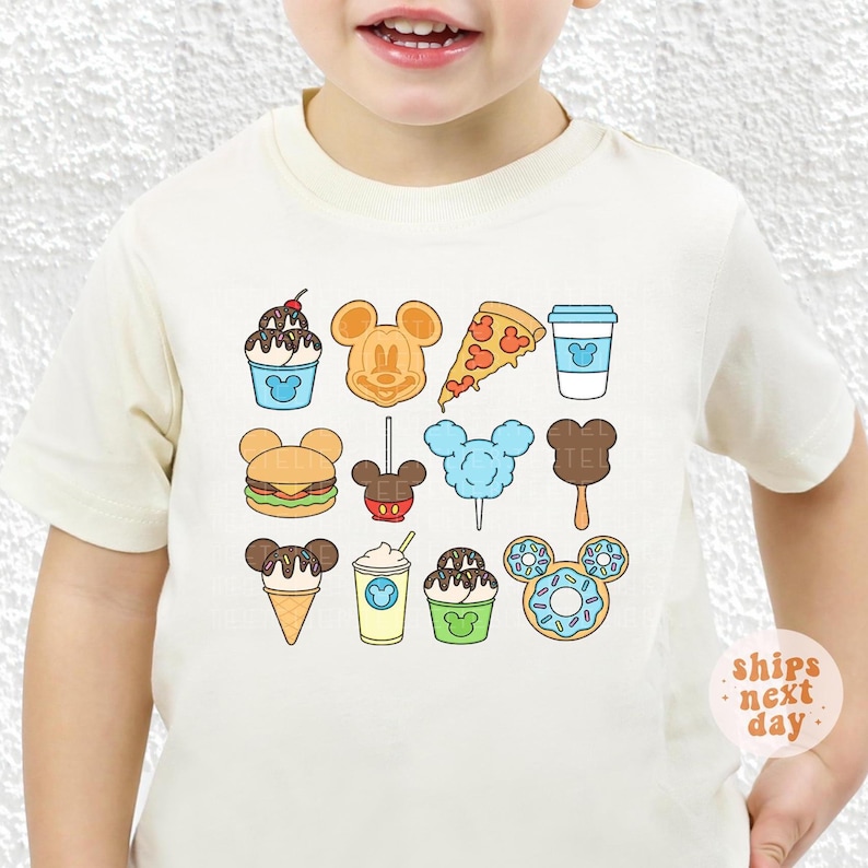 May include: A cream-colored short-sleeve t-shirt featuring a graphic print of various food items shaped like Mickey Mouse ears, including a waffle, pizza slice, burger, ice cream, cotton candy, and a donut, all arranged in a grid pattern.