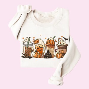 May include: A white sweatshirt with a graphic print of four pumpkin spice-themed drinks with ghosts, bats, and stars. The drinks are decorated with whipped cream, sprinkles, and pumpkin accents.