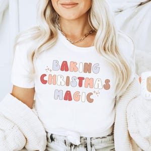 May include: White t-shirt with the text "BAKING CHRISTMAS MAGIC" in colorful retro-style lettering. The shirt has a crew neck and is made of a soft material. The design includes stars and a festive color palette.