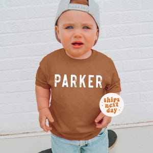 May include: A short-sleeved, solid rust-colored t-shirt featuring the name PARKER printed in large, white, distressed-style capital letters across the chest. The shirt is paired with light blue denim jeans and a grey baseball cap.