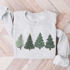 May include: A light gray crewneck sweatshirt featuring a graphic print of four stylized evergreen trees. The trees are depicted in varying shades of green with hand-drawn details, including small yellow ornaments and a star on one tree.