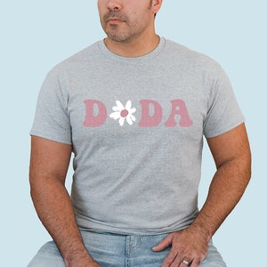 Puede incluir: Una camiseta gris con un gr&aacute;fico rosa "D*DA" con una margarita blanca.