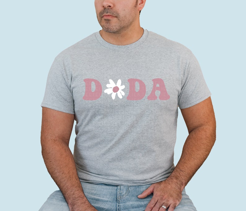 Puede incluir: Una camiseta gris con un gr&aacute;fico rosa "D*DA" con una margarita blanca.