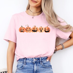 Pu&ograve; includere: T-shirt rosa chiaro con una fila di zucche di Halloween in stile acquerello in arancione e nero. Ogni zucca ha un volto intagliato unico. La maglietta &egrave; a girocollo.