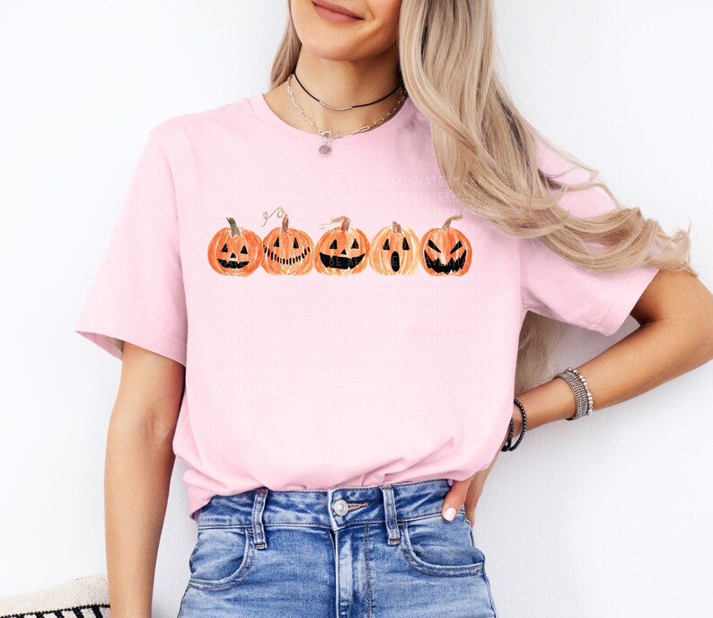 Pu&ograve; includere: T-shirt rosa chiaro con una fila di zucche di Halloween in stile acquerello in arancione e nero. Ogni zucca ha un volto intagliato unico. La maglietta &egrave; a girocollo.