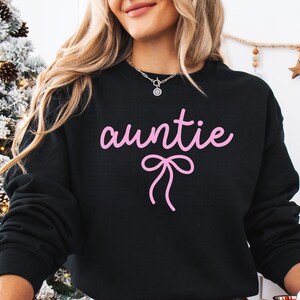 May include: Black sweatshirt with the word "auntie" in pink script and a pink bow graphic. The sweatshirt is a crew neck style. The image shows a close-up of the upper body.