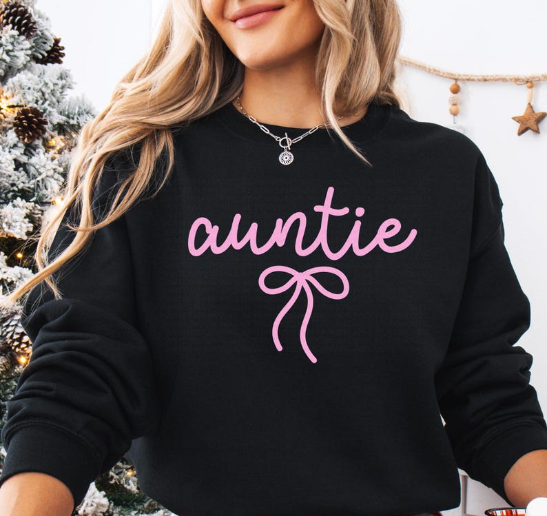 May include: Black sweatshirt with the word "auntie" in pink script and a pink bow graphic. The sweatshirt is a crew neck style. The image shows a close-up of the upper body.
