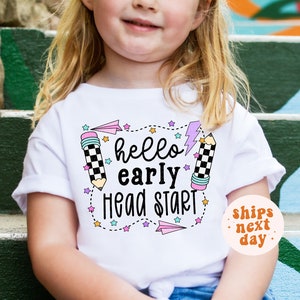 May include: White t-shirt with a colorful graphic that says "hello early head start" with pencils, stars, and paper airplanes. The shirt is being worn by a child.